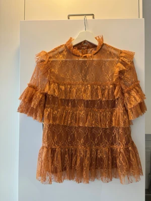 Orange spetsblus med volanger - Trendig orange blus i genomskinlig spets med volangdetaljer på både ärmar och kropp. Blusen har hög hals och trekvartsärm, samt ett romantiskt och lekfullt uttryck. Perfekt för dig som vill sticka ut med en unik och färgstark look.
