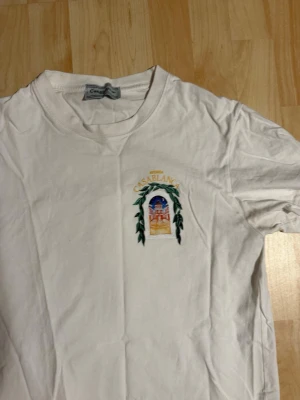 Vit Casablanca t-shirt med tryck - Säljer en vit t-shirt från Casablanca med färgglatt tryck fram och bak. På bröstet finns ett litet broderat motiv och på ryggen ett stort tryck med texten 'Avenida Casablanca' och ett konstnärligt motiv av en byggnad och gröna blad. Klassisk passform och rund hals.