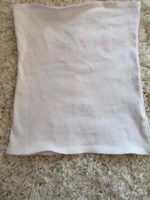 Vit ribbad tubtopp - Säljer en enkel och stilren vit ribbad tubtopp i storlek S. Toppen är axelbandslös och har en mjuk, stretchig känsla som sitter snyggt på kroppen. Perfekt att matcha med jeans eller kjol för en clean look.