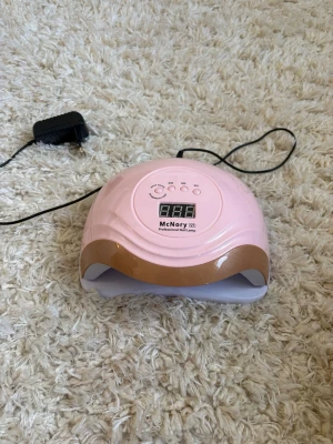 Rosa UV-lampa för naglar McNory - Elektrisk UV-lampa för naglar från McNory i rosa med gulddetalj. Lampan har digital display, flera timerinställningar och medföljande strömadapter. Perfekt för att härda gel- och shellacknaglar hemma.