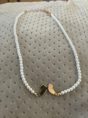 Pärlhalsband med guldhjärta - Stilrent halsband med vita pärlor och ett guldfärgat hjärthänge i mitten. Perfekt accessoar för att lyfta din outfit med en söt och trendig detalj. Kedjan har justerbar längd och pärlorna ger en klassisk känsla.