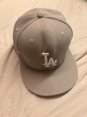 Grå LA keps med broderad logga - Snygg grå keps med rak skärm och broderad LA-logga framtill i vitt. På sidan finns ytterligare ett broderat motiv. Kepsen har klassisk snapback-form och är tillverkad i ett tåligt tygmaterial. Perfekt för dig som vill ha en stilren och sportig look.