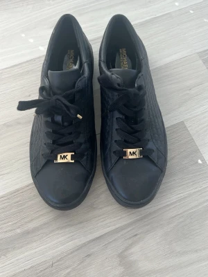Svarta Michael Kors sneakers - Snygga svarta sneakers från Michael Kors med guldfärgade MK-detaljer på snörningen. Skorna har en låg profil och är gjorda i ett läderliknande material med diskret mönster på sidorna. Perfekta för dig som gillar stilrena och trendiga skor. 