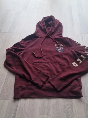 Vinröd hoodie från Hollister - Snygg vinröd hoodie från Hollister med tryck på bröstet och text längs ena ärmen. Klassisk känguruficka framtill och justerbar huva med dragsko. Perfekt för dig som gillar en avslappnad och trendig stil.