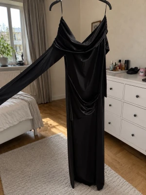 Svart långklänning med drapering - Elegant svart långklänning i glansigt material med draperad omlott-ringning och långa ärmar. Klänningen har en smickrande passform och faller snyggt längs kroppen. Perfekt för dig som vill sticka ut med en stilren look. Helt ny och väldigt stretchig 