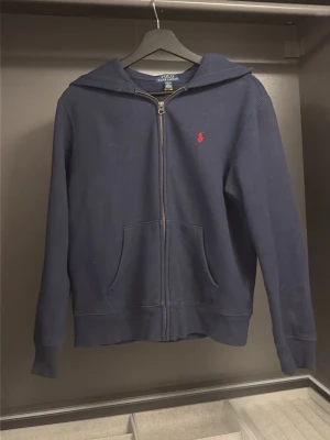 Marinblå hoodie från Polo Ralph Lauren - Klassisk marinblå hoodie med dragkedja från Polo Ralph Lauren. Har huva, två fickor framtill och den ikoniska röda logotypen broderad på bröstet. Tillverkad i mjukt bomullsmaterial som känns skönt mot huden. Perfekt för en avslappnad och stilren look.