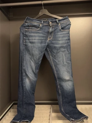 Blå jeans från Replay - Snygga blå jeans från Replay med klassisk femficksmodell och raka ben. Jeansen har slitningar och ljusare partier för en trendig look. Tillverkade i denim med kontrastsömmar och knappgylf. Perfekta för dig som gillar en avslappnad men stilren stil.