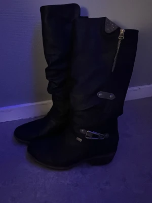 Svarta höga boots med spänne - Säljer ett par svarta höga boots med dekorativt spänne och dragkedja på sidan. Stövlarna har en liten klack och detaljer i metall, samt en något rynkad design på skaftet. Perfekta för dig som vill ha en edgy och stilren look.