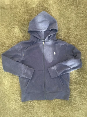 Polo Ralph Lauren Hoodie - Säljer en klassisk zip hoodie från Polo Ralph Lauren i marinblå färg. Broderad vit Polo-logga på bröstet och hel dragkedja framtill. Mjuk och bekväm med justerbar huva och två framfickor – en tidlös hoodie som passar till allt.  • Märke: Polo Ralph Lauren • Färg: Marinblå • Modell: Zip Hoodie • Skick: Bra använt skick, utan större defekter. • Perfekt till vardags och enkel att matcha med både jeans och mjukisbyxor.  📦 Skickas snabbt och väl paketerat.. Skicka gärna ett meddelande för fler bilder