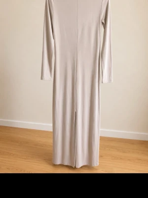 Beige långärmad långklänning - En stilren beige långklänning med lång ärm och rak passform. Klänningen har en enkel design utan mönster och en diskret slits där bak. Perfekt för dig som gillar minimalistisk stil och mjuka material. Klänningen är väldigt stretchig