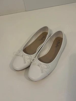Vita ballerinaskor med rosett - Stilrena vita ballerinaskor från Duffy med rund tå och söt rosettdetalj framtill. Skorna har en klassisk låg klack och är tillverkade i ett slätt material som ger en clean look. Skorna är använda 1 gång och ser helt nya ut! De är i storlek 39💕