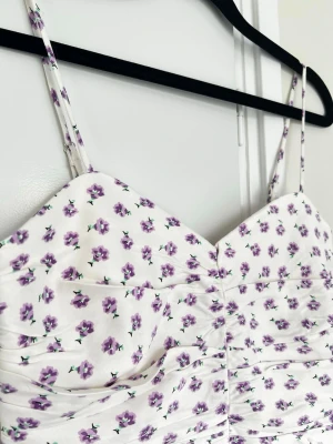 Satin-topp med lika blommor (nyskick) - En croppad, vit topp i satinmaterial med små lila blommor, från Zara. Rynkad i sidorna och smock i ryggen. Dragkedja i sidan. Aldrig använd. Ej stretch.
