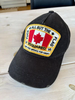 Svart Dsquared2 keps med patch - Säljer en svart keps från Dsquared2 med stor patch framtill med kanadensisk flagga och texten 'All But The Flag'. Justerbar rem bak med gul brodyr 'Dean & Dan Born In Canada'. Klassisk böjd skärm och slitna detaljer för en cool look.