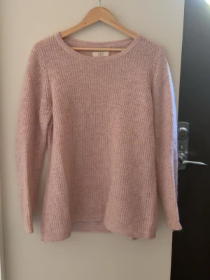 Ljusrosa stickad tröja - Mysig ljusrosa stickad tröja med rund halsringning och långa ärmar. Tröjan har en enkel, rak passform och är tillverkad i ett mjukt, fluffigt material som känns skönt mot huden. Perfekt för kyliga dagar och enkel att matcha med olika outfits.