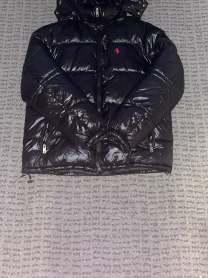 Svart dunjacka med huva från Polo Ralph Lauren - Säljer en svart, glansig dunjacka från Polo Ralph Lauren med huva och dragkedja framtill. Jackan har två sidofickor med dragkedja och den klassiska röda loggan på bröstet. Perfekt för kalla vinterdagar och riktigt snygg streetstil.