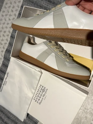 Maison Margiela Replica vita sneakers - Snygga Maison Margiela Replica sneakers i vitt och ljusgrått med beige skosnören och klassisk gummisula. Ovandelen är i en mix av läder och mocka för en clean och stilren look. Perfekta för dig som gillar minimalistisk och trendig stil. Helt oanvända, pris går att diskuteras vid snabb köp!🫡