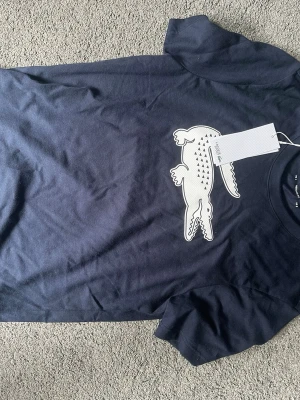 Marinblå Lacoste Sport t-shirt - Snygg marinblå t-shirt från Lacoste Sport med stor vit krokodil-logga på bröstet. Klassisk rund hals och korta ärmar. Tillverkad i mjuk bomull för skön känsla och perfekt passform. Perfekt för dig som gillar stilrena och sportiga plagg.