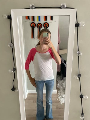 Vit och rosa baseball t-shirt - Säljer en vit t-shirt med rosa trekvartsärmar i klassisk baseballstil. Tröjan har rund halsringning och är lite längre baktill. Perfekt för en avslappnad och sportig look.