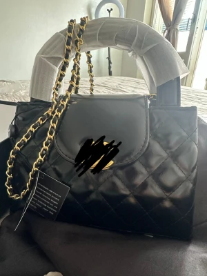Svart quiltad handväska med kedja - Snygg svart handväska i quiltat mönster med guldfärgad kedjerem och topphandtag. Väskan har lockstängning och en elegant, stilren look som passar till många outfits. Materialet ser ut att vara konstläder och kedjan ger en lyxig känsla.
