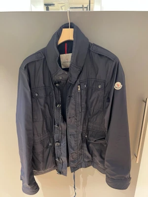 Navy/Blå Field Jacka från Moncler - Snygg blå vindjacka från Moncler med hög krage, dragkedja och knappar framtill. Jackan har flera fickor med lock och dragkedja samt Moncler-logga på ärmen. Tillverkad i ett lätt och tåligt material som passar perfekt till blåsiga dagar. Jackan har även en luva. Perfekt skick och inga defekter!