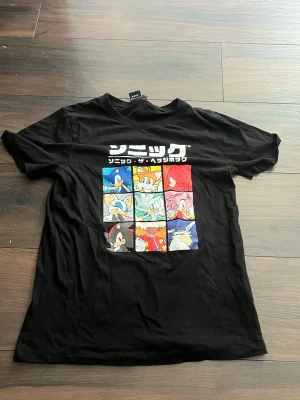 Svart Sonic the Hedgehog t-shirt - Säljer en svart t-shirt med färgglatt tryck av Sonic the Hedgehog och flera karaktärer från spelet på bröstet. T-shirten har korta ärmar och rund hals. Perfekt för dig som gillar retrospel och vill ha en unik look.