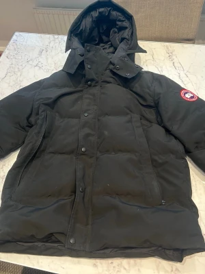 Svart dunjacka från Canada Goose - Säljer en svart dunjacka från Canada Goose med huva och klassisk logga på ärmen. Jackan har dragkedja och knappar framtill samt två stora fickor. Perfekt för kalla vinterdagar tack vare det tjocka dunfodret.