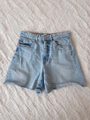 Ljusblå jeansshorts från Kappahl - Säljer ett par ljusblå jeansshorts från Kappahl med hög midja och rå kant nertill. Klassisk femficksmodell med knapp och dragkedja framtill. Perfekta för varma dagar och enkel att matcha med olika toppar.