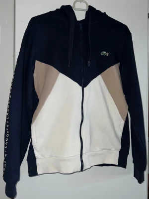 Lacoste hoodie med dragkedja - Snygg hoodie från Lacoste i marinblått, beige och vitt med dragkedja framtill. Har huva med snören och Lacoste-logga på bröstet. Ärmarna har texten 'LACOSTE' längs sidan. Perfekt för en sportig och avslappnad stil.