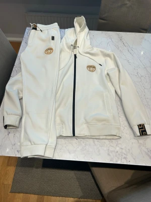 Vitt set med huva och guldmönster - Snyggt vitt set med zip-hoodie och matchande byxor. Setet har guldfärgade detaljer och emblem på både jacka och byxa samt dekorativa band med svart och guldmönster på huvan och midjan. Mjukt material och avslappnad passform, perfekt för en trendig look.