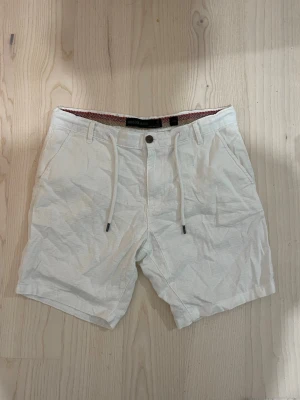 Vita linneshorts med snörning - Snygga vita shorts i linne med snörning i midjan och knappstängning. Klassisk passform med sidofickor fram och bakfickor med knapp. Perfekta för varma dagar och har bälteshällor för extra stil.