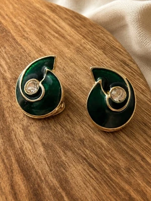 Gröna swirl örhängen med kristall - Unika örhängen i swirl-form med djupgrön emalj och guldfärgade kanter. I mitten sitter en klar kristall som ger extra bling. Clip-on-modell, perfekta för dig som vill sticka ut med coola detaljer och retrovibbar.