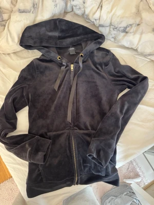 Mjuk svart hoodie från Lindex - Säljer en svart hoodie från Lindex i mjuk velour med dragkedja framtill, stora fickor och huva med breda snören. Guldiga detaljer vid snörhålen ger en snygg touch. Perfekt för dig som gillar en chill och bekväm stil.