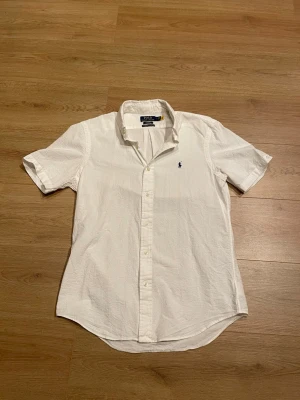 Vit kortärmad skjorta Polo Ralph Lauren - Klassisk vit kortärmad skjorta från Polo Ralph Lauren med broderad logga på bröstet. Skjortan har knappar framtill och är tillverkad i bomull med en lätt strukturerad yta. Perfekt för en clean och stilren look. Köpt ny av mig och är inte använd 