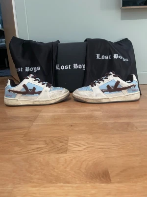 Lost Boys blå och vita sneakers - Säljer ett par sneakers från Lost Boys i vitt och ljusblått med bruna detaljer och snörning. Skorna har en låg silhuett och en unik design med kontrasterande färgpaneler och logga på sidan. Perfekta för dig som gillar streetwear och vill sticka ut.