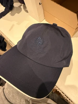 Mörkblå keps från LP - Snygg mörkblå keps från LP med broderad logga och text framtill. Klassisk böjd skärm med vit kantdetalj och justerbar passform bak. Tillverkad i ett lätt och bekvämt material som passar perfekt till streetwear eller casual outfits.