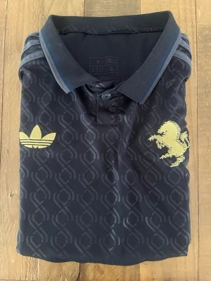 Juventus 2024/2025 Mörkblå  - Mörkblå pikétröja från Adidas i samarbete med Juventus. Tröjan har diskret mönster i tyget, guldfärgade Adidas-logga och Juventus-emblem på bröstet samt guldfärgad 'JUVENTUS' text i nacken. Klassisk krage och korta ärmar. Perfekt för fotbollsfans.