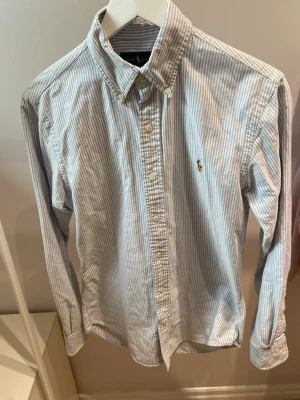 Randig skjorta med knappar - Klassisk skjorta med smala blå och vita ränder, långärmad och med knappar hela vägen framtill. Skjortan har button-down krage och en liten broderad logga på bröstet. Perfekt för en stilren och avslappnad look.har tyvärr fått et litet hål+ en liten fläck(bild på) men knappt något som syns 