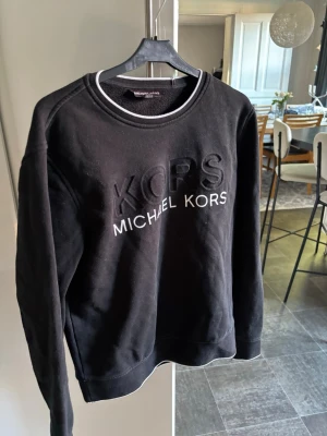 Svart sweatshirt från Michael Kors - Snygg svart sweatshirt från Michael Kors med broderad logga framtill. Tröjan har rund halsringning med vit detalj och långa ärmar. Perfekt för en stilren och avslappnad look.
