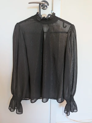 Svart transparent blus med volang - Snygg svart transparent blus med subtilt glittrande ränder. Blusen har hög krage med volangdetalj och långa ärmar med volang vid ärmslut. Perfekt för dig som gillar en trendig och elegant look.