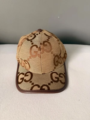 Beige keps med GG-mönster från Gucci - Snygg beige keps från Gucci med klassiskt GG-mönster i brunt över hela kepsen. Kepsen har bruna detaljer och justerbart band bak i läder. Tillverkad i Italien och perfekt för dig som vill ha en exklusiv och trendig accessoar.