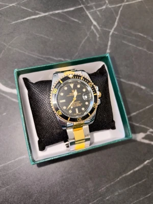Rolex Submariner silver/guld klocka - Snygg Rolex Submariner klocka i silver och guld med svart urtavla och gulddetaljer. Armbandet är i metall med tvåfärgad design och klassiskt Rolex-spänne. Urtavlan har tydliga index och datumfönster. Perfekt accessoar för dig som gillar exklusiv stil.
