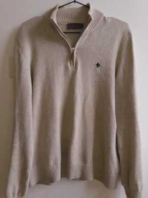 Morris Beige stickad tröja  - Beige stickad tröja med hög krage och halv dragkedja framtill. Tröjan har ribbade muddar vid ärmslut och nederkant samt en liten broderad logga på bröstet. Perfekt för lager på lager och enkel att matcha.
