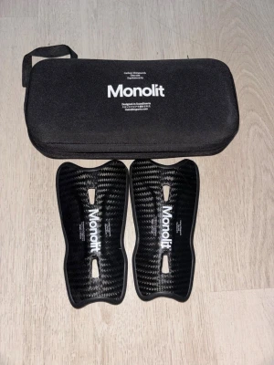 Monolit benskydd i kolfiber - Säljer ett par svarta benskydd från Monolit i kolfiber med snygg, modern design. De är lätta och har en ergonomisk form för bästa passform. Kommer med ett praktiskt svart fodral med dragkedja och handtag. Perfekta för dig som vill ha skydd med stil på planen.