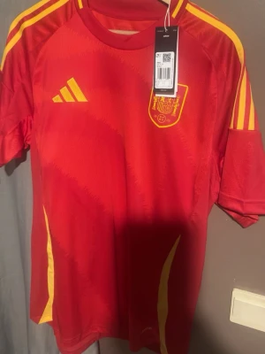 Spaniens matchtröja Adidas röd/gul 2024 - Säljer en officiell röd matchtröja från Spaniens landslag, tillverkad av Adidas. Tröjan har gula detaljer, tre ränder på axlarna och Spaniens klubbmärke på bröstet. Tillverkad i lätt och ventilerande material, perfekt för fotboll eller träning.