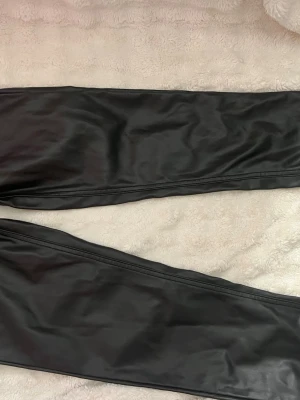 Svarta leggings i fuskskinn från 157 - Snygga svarta leggings i fuskskinn från 157. De har en tight passform och glansig finish som ger en cool look. Perfekta att styla med oversized tröjor eller crop tops. Materialet är mjukt och stretchigt för extra komfort.