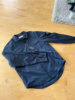 Mörkblå overshirt från Stone Island - Snygg mörkblå overshirt från Stone Island med dragkedja framtill, bröstficka och klassisk patch på ärmen. Tillverkad i ett lätt, slitstarkt material som ger en clean och modern look. Perfekt att bära över en t-shirt för en avslappnad stil.
