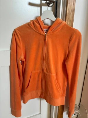 Orange velour hoodie Juicy Couture - Säljer en orange velour hoodie från Juicy Couture med dragkedja framtill och fickor. Baksidan har glittriga detaljer och texten 'Juicy Couture'. Storlek M men passar även S
