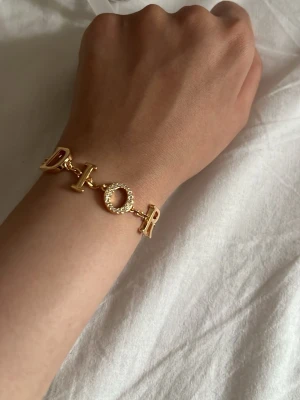 Guldarmband med pärldetaljer oxh paketering - Snyggt guldarmband där O:et är dekorerat med små pärlor. Armbandet har en elegant och modern design som verkligen sticker ut. Perfekt accessoar för dig som vill ha något exklusivt och trendigt.
