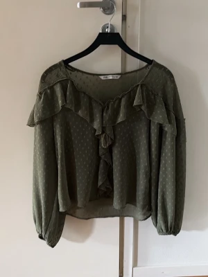 Blus från Zara - Snygg mörkgrön blus med volangdetaljer längs ringningen och framtill. Blusen har diskret prickmönster, v-ringning och långa ärmar med resår vid handlederna. Tillverkad i ett lätt, transparent tyg som ger en luftig känsla. Har köpt den secondhand och den var så lite sliten på volangen i ryggen men inget man märker av. 