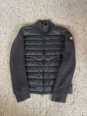 Moncler cardigan  - Snygg svart cardiganjacka från Moncler med quiltad front och stickade ärmar. Jackan har hel dragkedja framtill och en liten Moncler-logga på ärmen. Perfekt för lager-på-lager och en clean look.